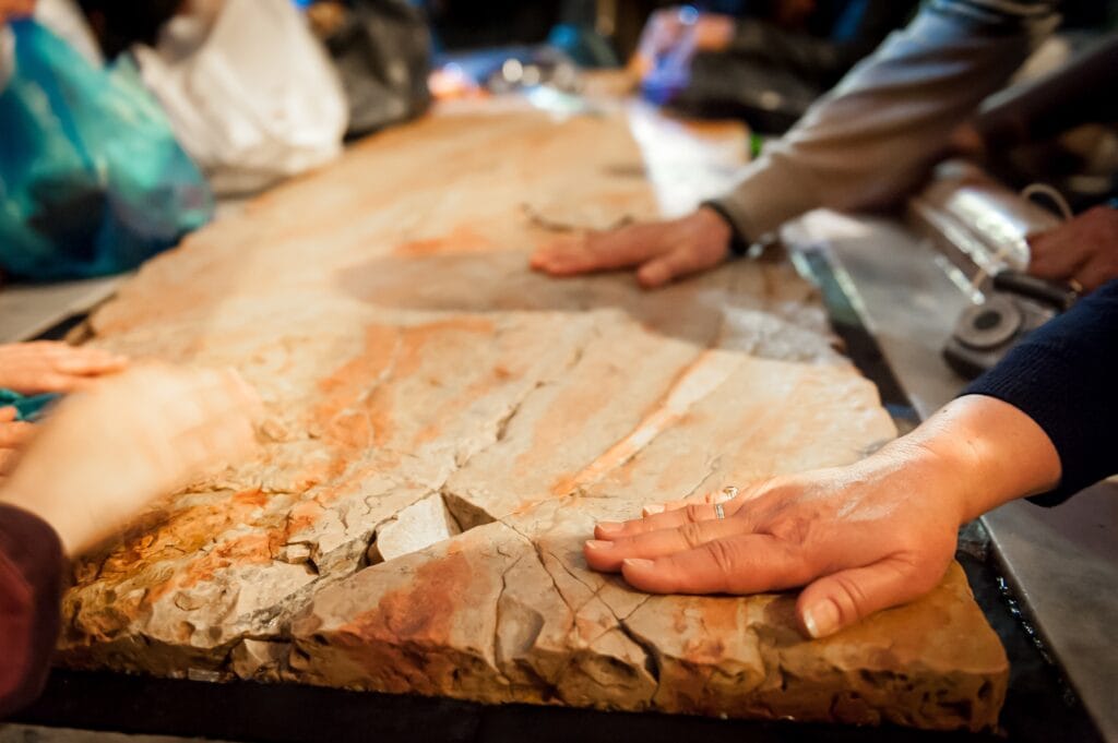 Several people touch a large, flat, textured stone slab with both hands. The stone has orange and beige streaks, and the photo is taken indoors under warm lighting—an impressive showcase of craftsmanship by Bay Area Stone Experts.